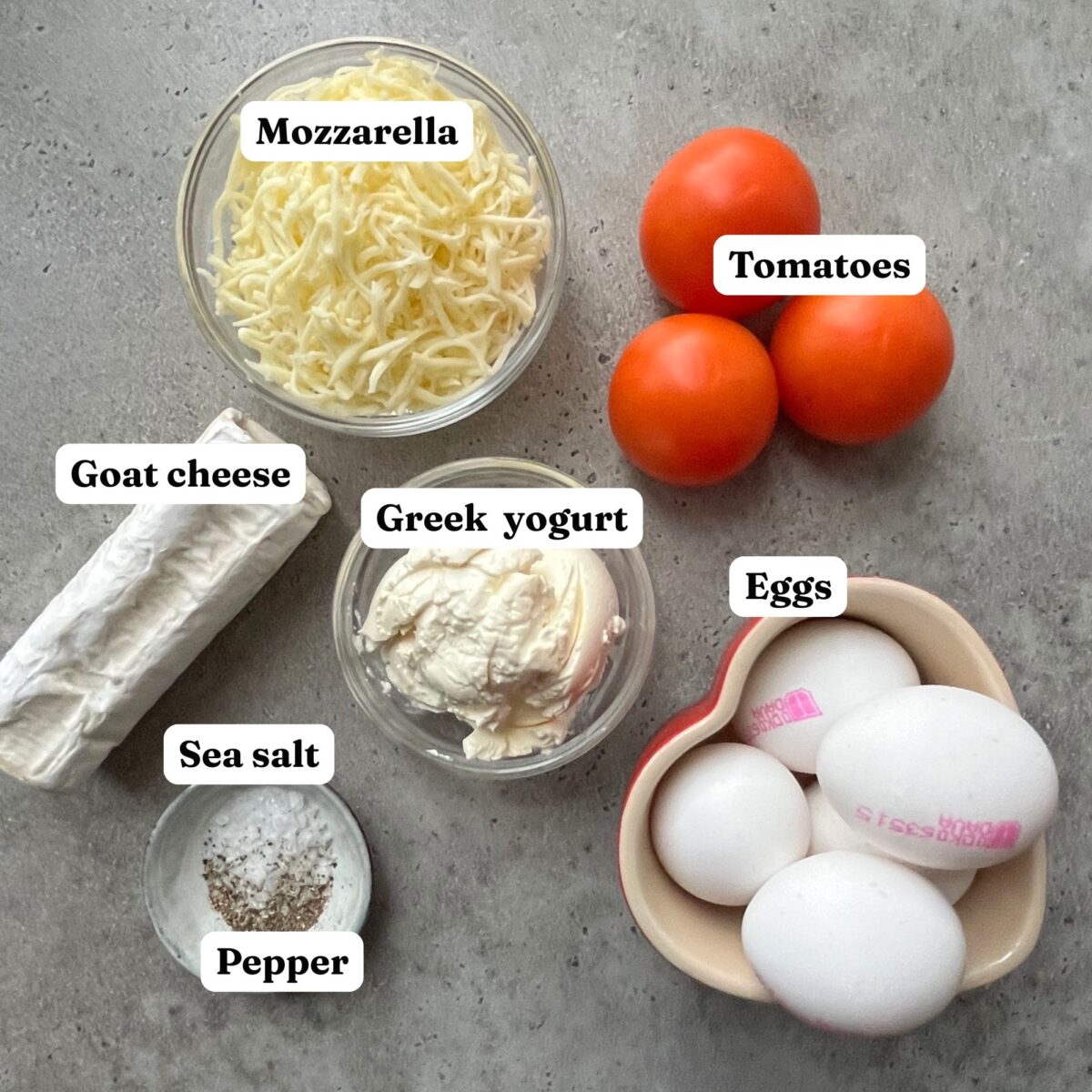 Shredded mozzarella, whole tomatoes, goat cheese, Greek yogurt, and four eggs in a bowl come together as the perfect ingredients for a flavorful Goat Cheese Quiche, with small dishes of sea salt and pepper nearby.