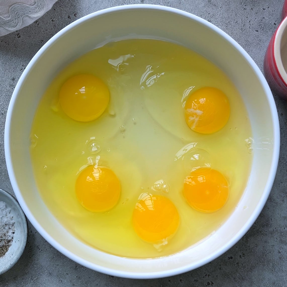 A white bowl containing raw eggs with intact yellow yolks and clear egg whites on a gray countertop.