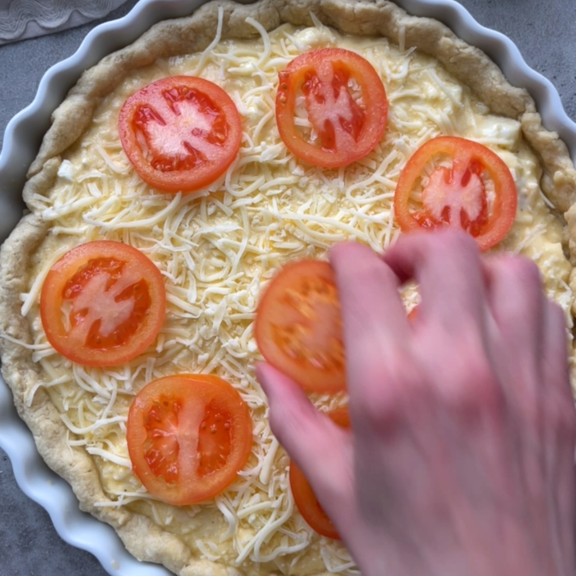 A hand places tomato slices on an unbaked cheese-covered quiche in a round baking dish.