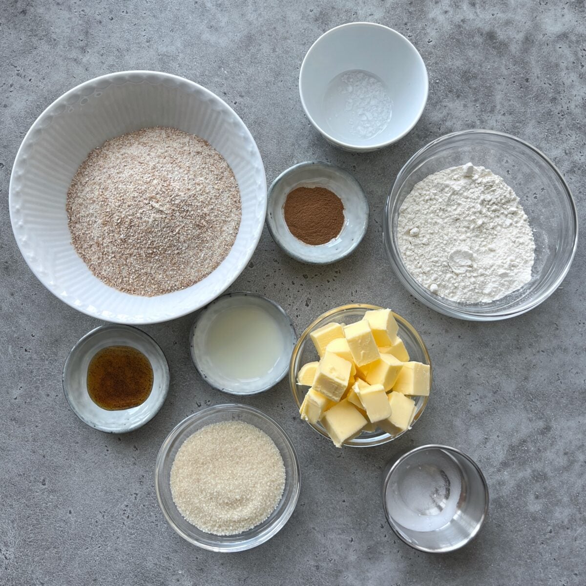 Nine bowls containing graham flour, plain flour, butter cubes, sugar, milk, vanilla extract, cinnamon, baking soda, and salt. 