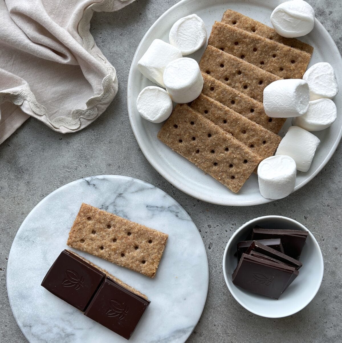A plate with homemade graham crackers and marshmallows, a marble dish with a graham cracker and chocolate squares, and a bowl of chocolate pieces on a gray surface.