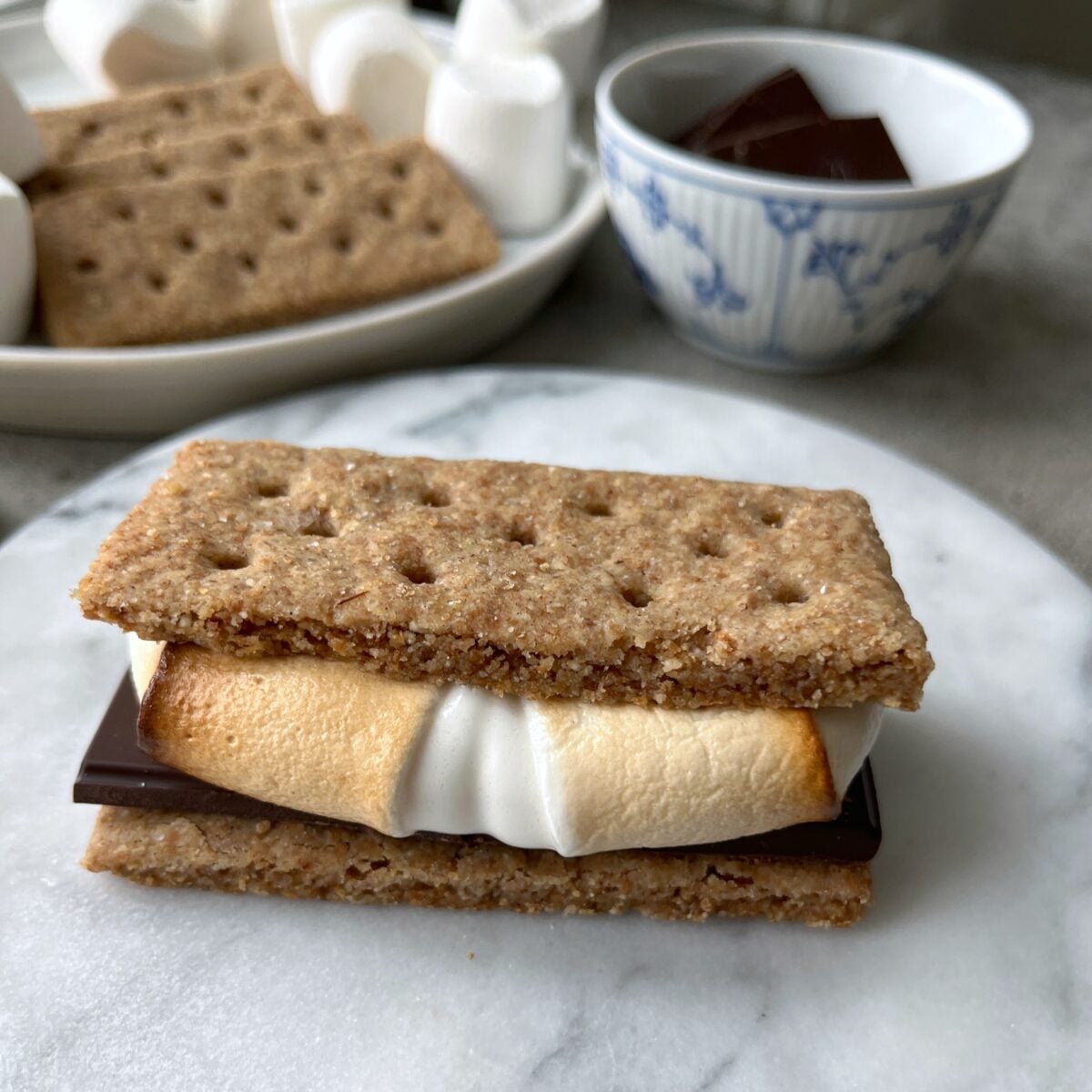 A s'more made with homemade graham crackers, a toasted marshmallow, and chocolate sits on a marble surface, with extra graham crackers, marshmallows, and chocolate in the background.
