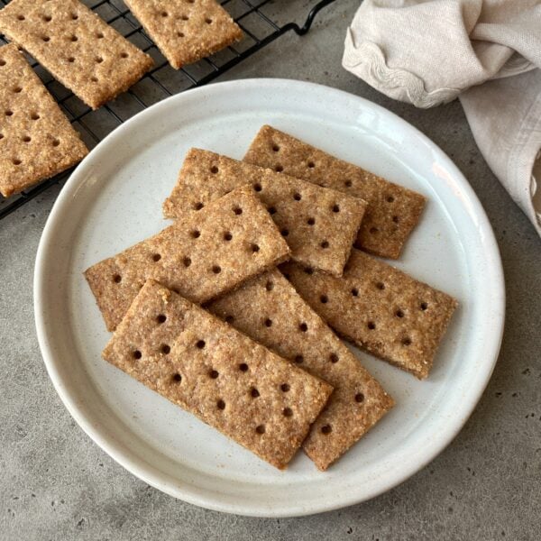 A plate with several rectangular homemade graham crackers, each with rows of small holes, on a gray surface. More homemade graham crackers are cooling on a rack in the background.