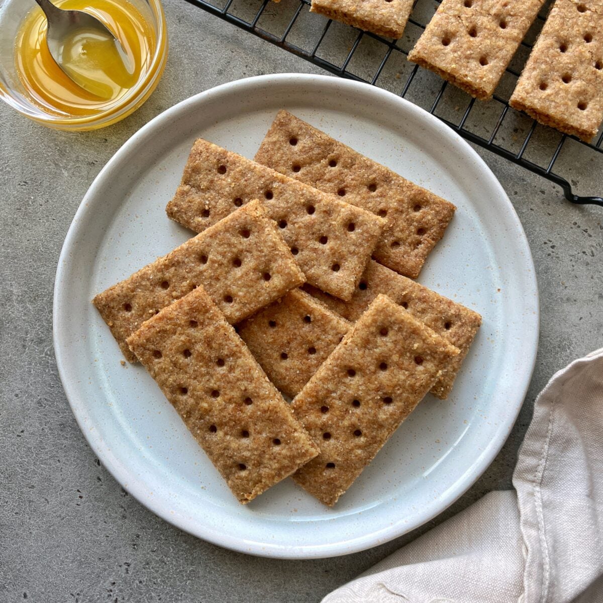 A plate of rectangular, Homemade Graham Crackers with uniform holes is shown next to a small bowl of honey and a cooling rack with more crackers.
