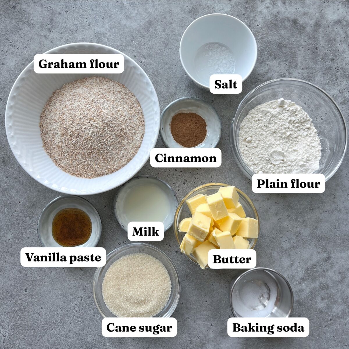 Nine bowls containing graham flour, plain flour, butter cubes, sugar, milk, vanilla paste, cinnamon, baking soda, and salt.