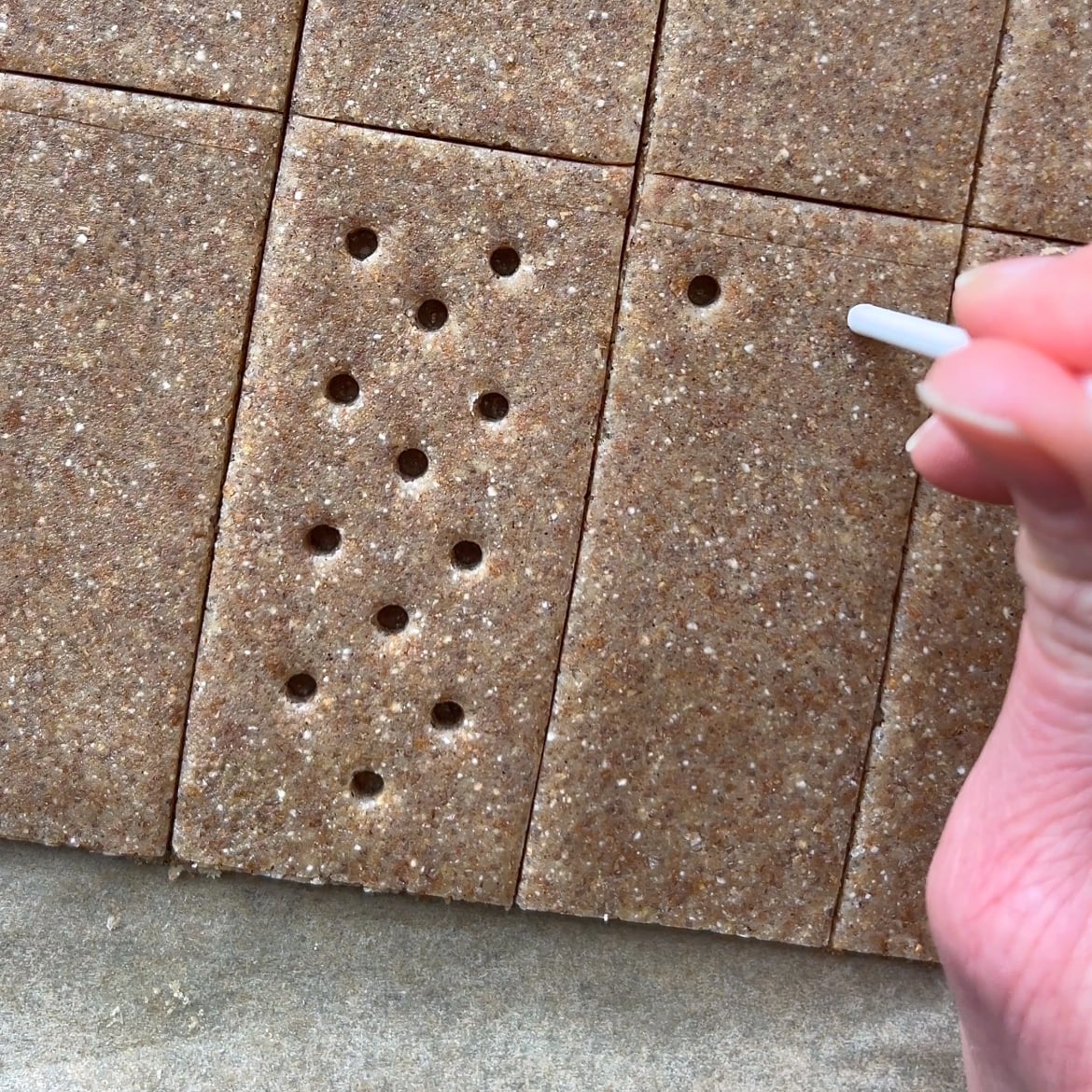 A hand uses a small lolly stick to poke holes in rectangular pieces of homemade graham cracker dough.