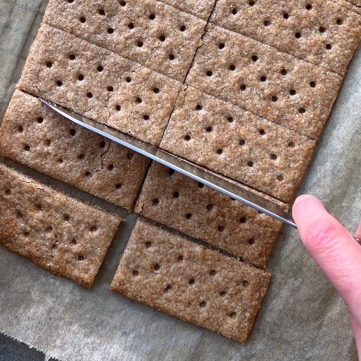 A hand uses a knife to cut rectangular Homemade Graham Crackers with small holes on top, placed on parchment paper.