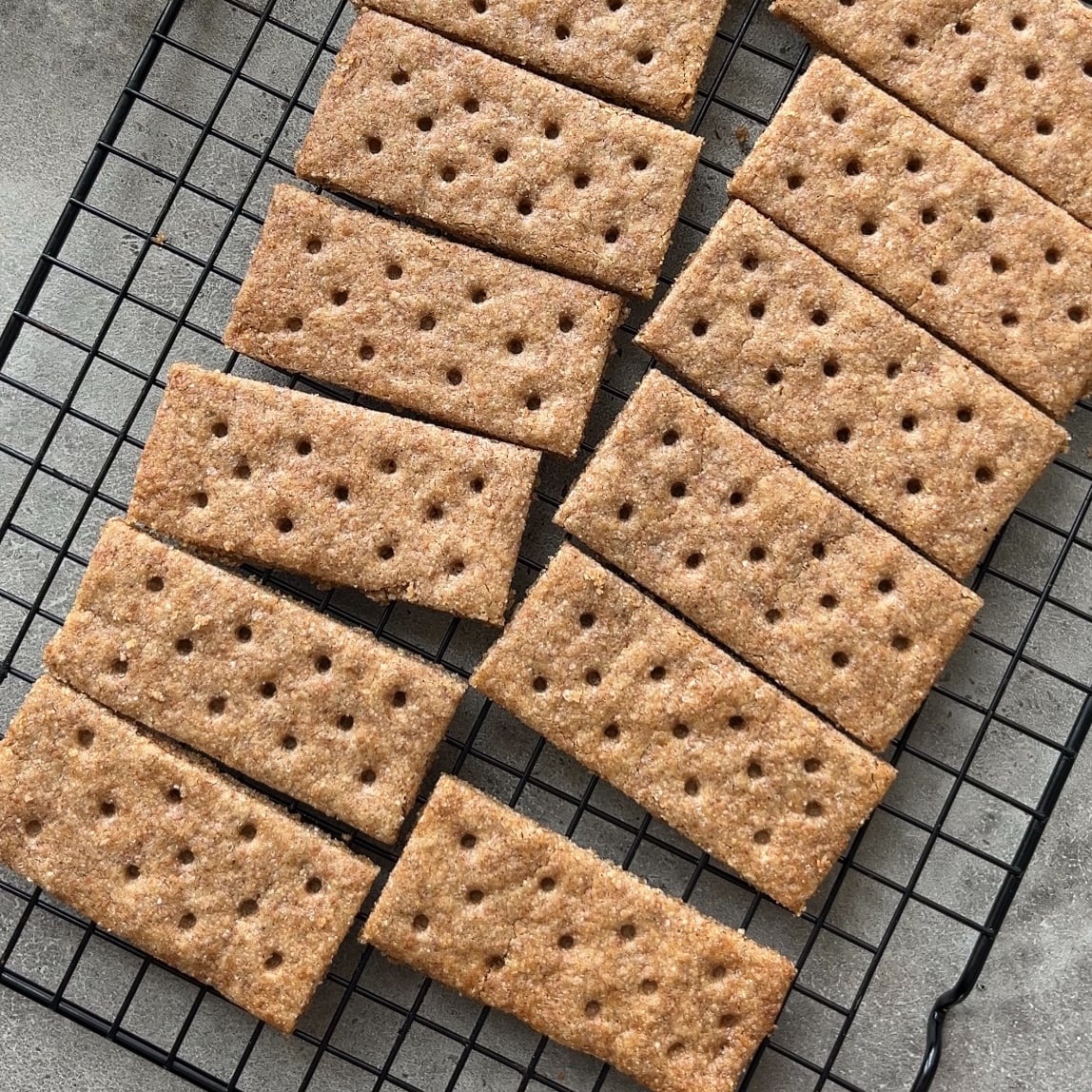 Homemade Graham Crackers, rectangular and brown with evenly spaced holes, are cooling on a black wire rack set atop a gray surface.