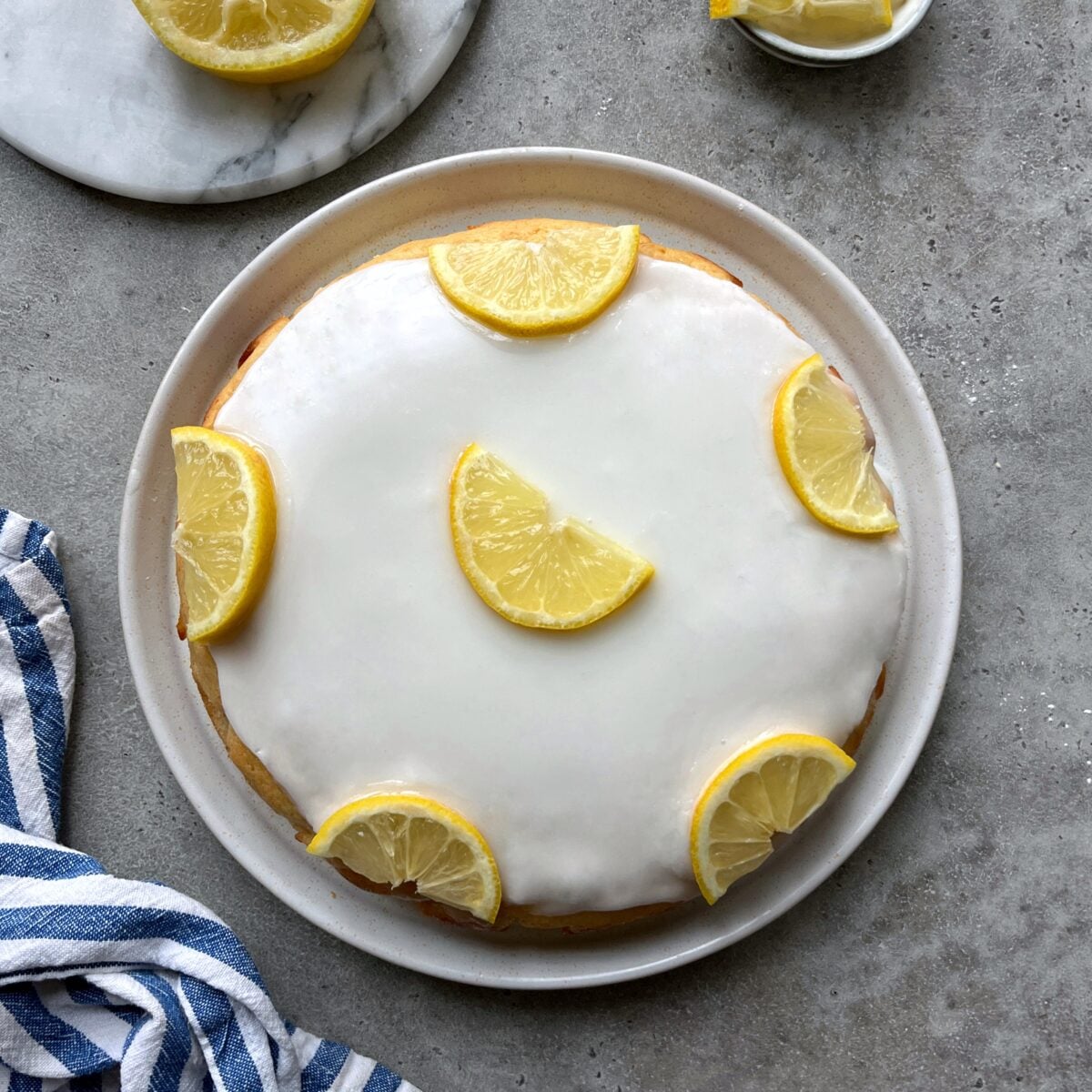 A round cake with white icing, topped with six lemon slices, sits on a plate. A striped towel and extra lemon slices are nearby on a gray surface.