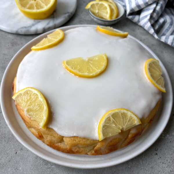 Round lemon cake with white icing, topped with six halved lemon slices, displayed on a white plate. A sliced lemon and cloth napkin are in the background.