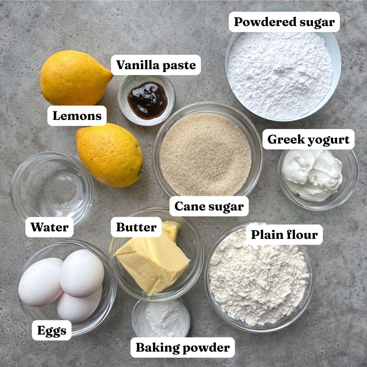 Various baking ingredients for Lemon Cake with Glaze, including lemons, vanilla paste, powdered sugar, cane sugar, Greek yogurt, water, butter, plain flour, baking powder, and eggs are arranged on a gray surface.