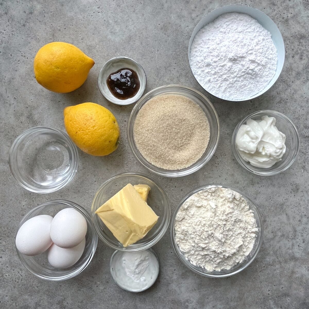 Ingredients for baking displayed on a gray surface, including lemons, eggs, butter, sugar, flour, powdered sugar, yogurt, vanilla paste, baking powder, and a small bowl of water.
