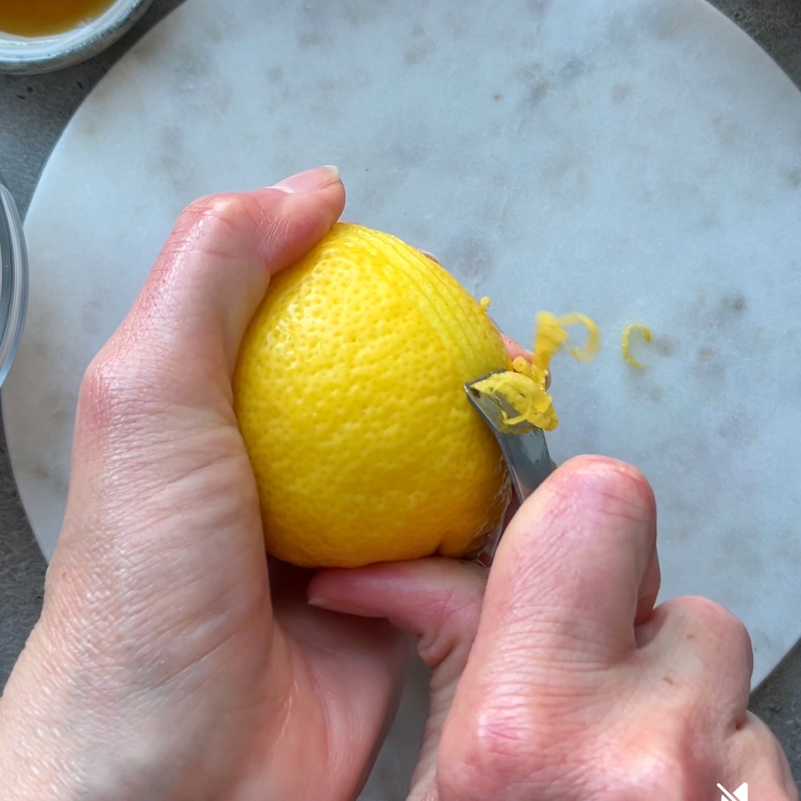 A hand holding a lemon while using a small knife to peel off thin strips of lemon zest over a round marble surface.