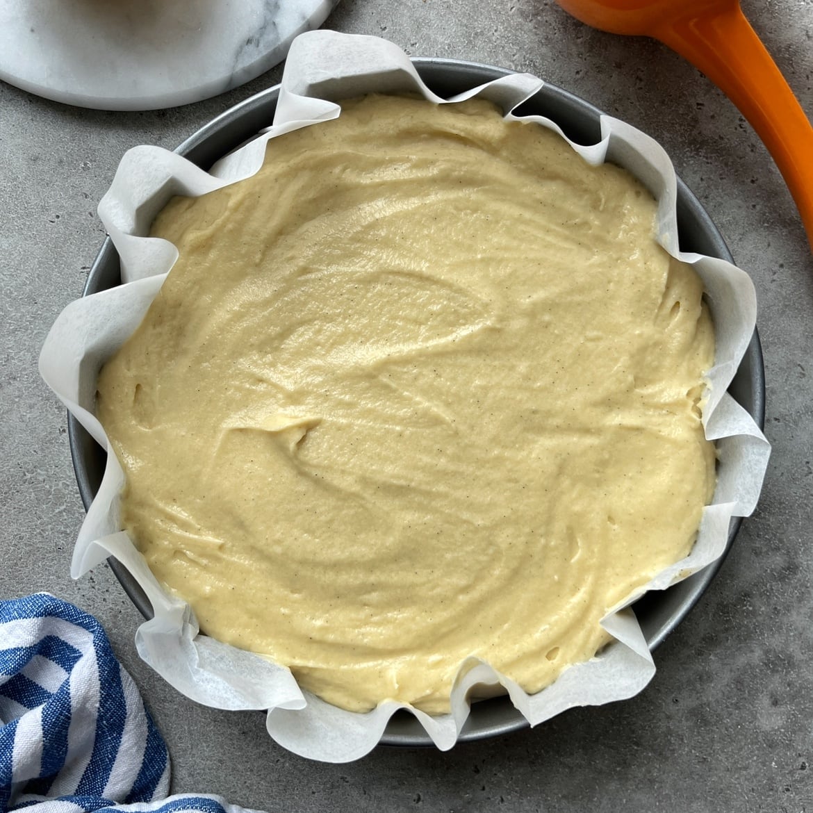 A round cake pan lined with parchment paper is filled with smooth lemon cake batter.