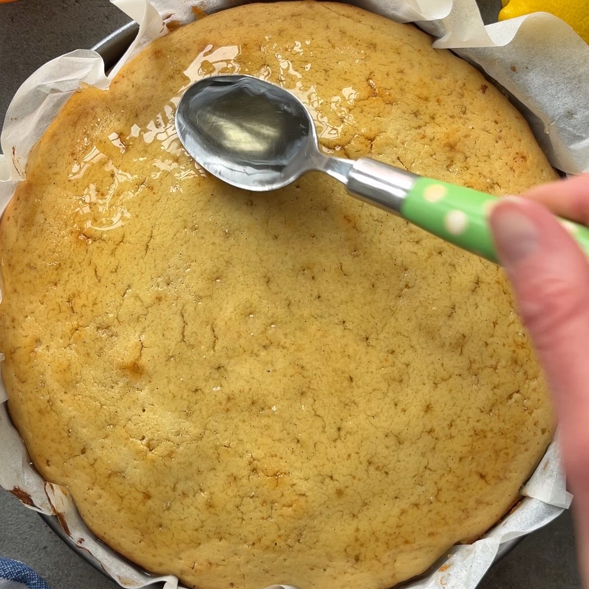 A hand holds a spoon, spreading syrup over a round, golden-brown Lemon Cake with Glaze in a parchment-lined pan.