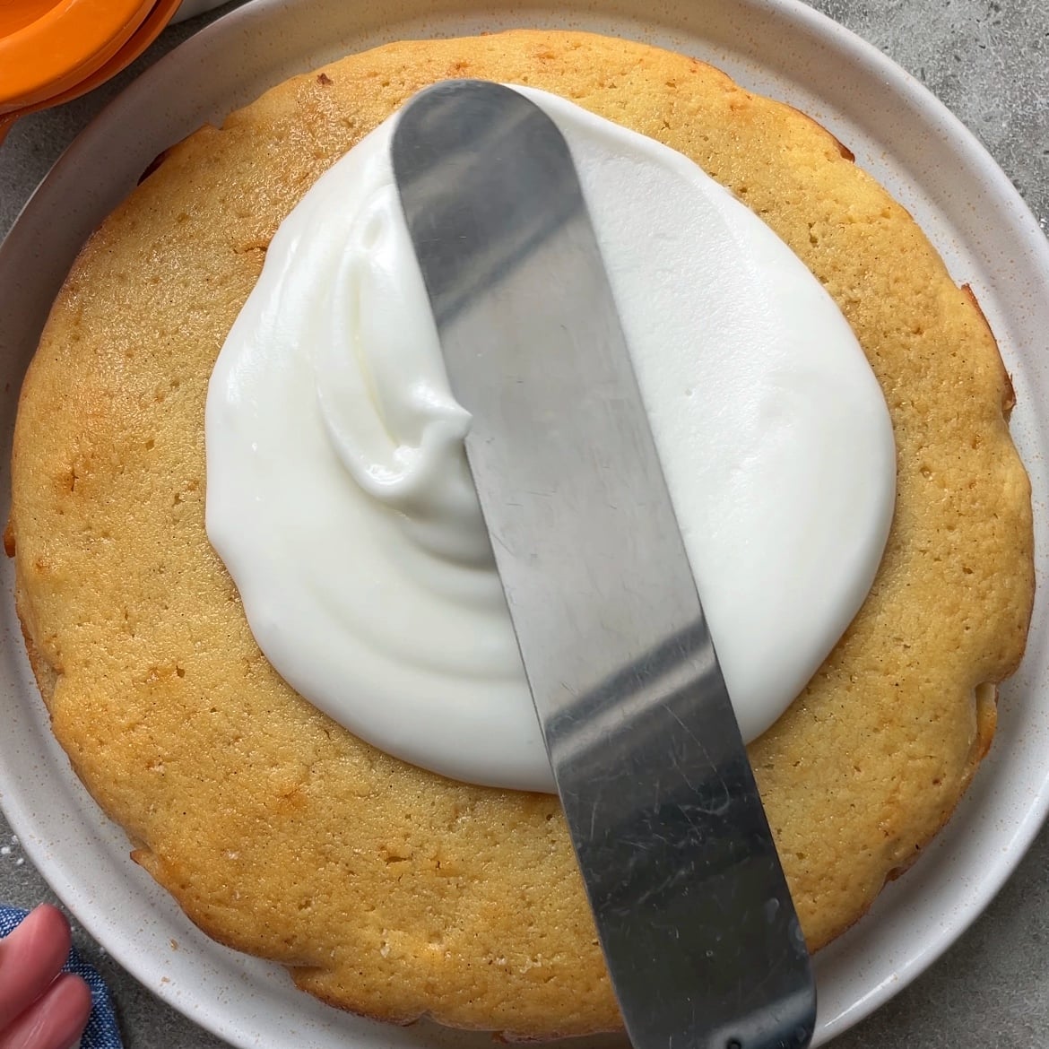 An offset spatula spreads white frosting over a round, unfrosted Lemon Cake with Glaze on a plate.
