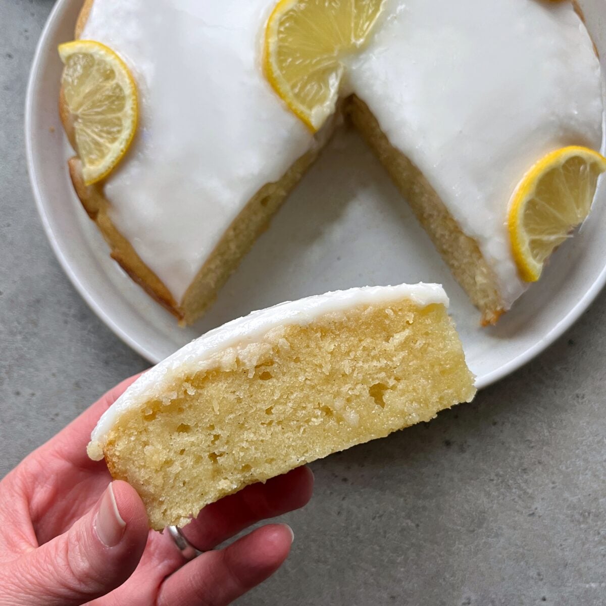 A hand holds a slice of lemon cake with white icing, while the rest of the cake, topped with lemon slices, sits on a white plate in the background.