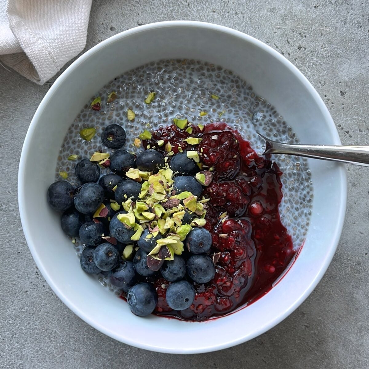 A bowl of chia pudding and Greek yogurt topped with blueberries, chopped pistachios, and a berry sauce, with a spoon resting inside the bowl.
