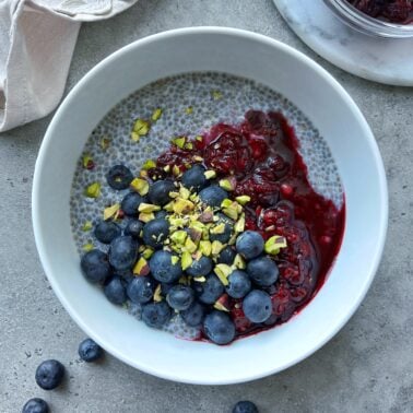 A white bowl of chia pudding and Greek yogurt topped with fresh blueberries, berry compote, and chopped pistachios, placed on a gray surface.