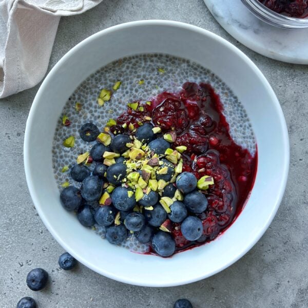 A white bowl of chia pudding and Greek yogurt topped with fresh blueberries, berry compote, and chopped pistachios, placed on a gray surface.