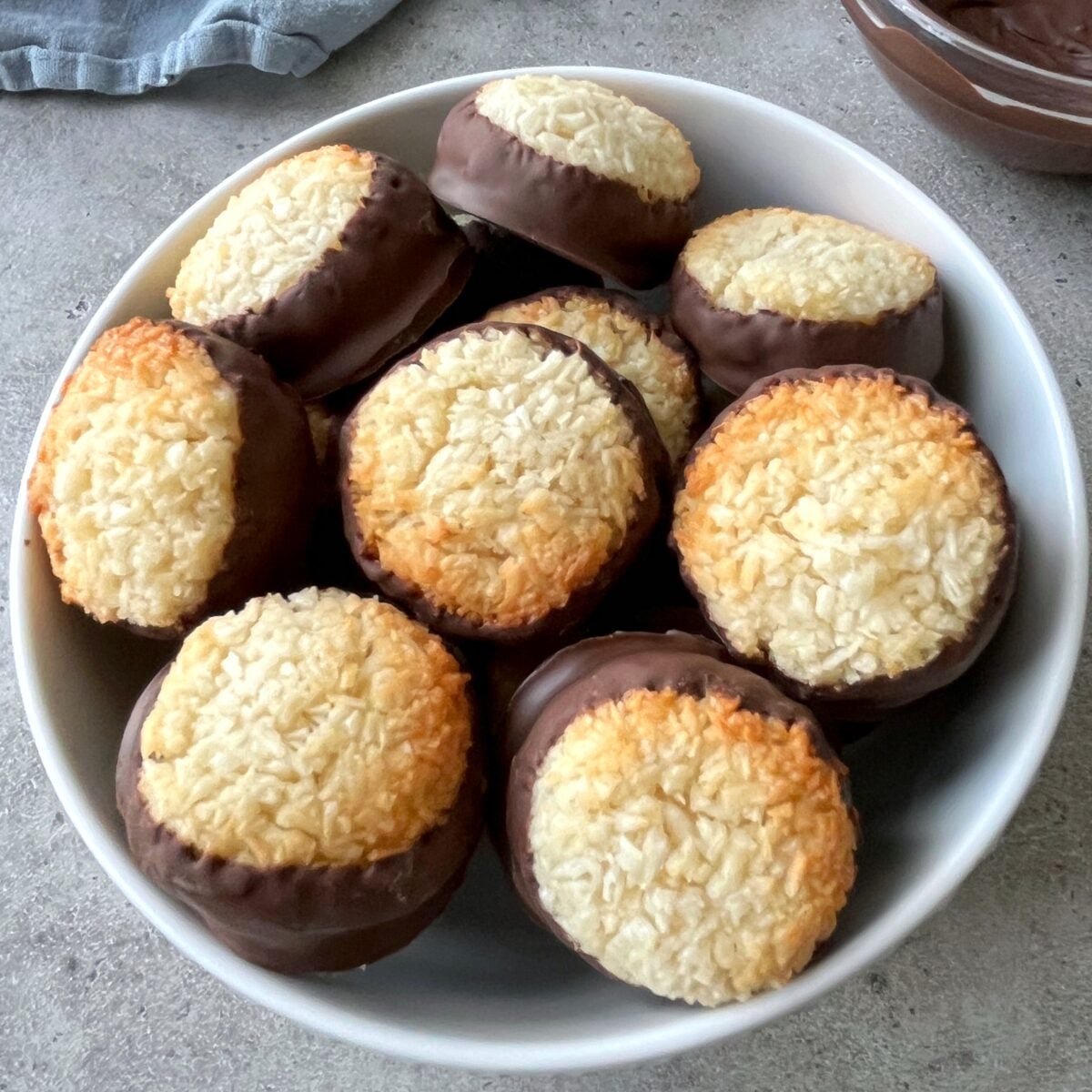 A white bowl filled with round coconut cookies, each partially dipped in chocolate, on a gray surface.