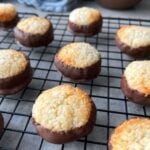 Round coconut cookies, shaped as macaroons and partially dipped in chocolate, are cooling on a black wire rack.