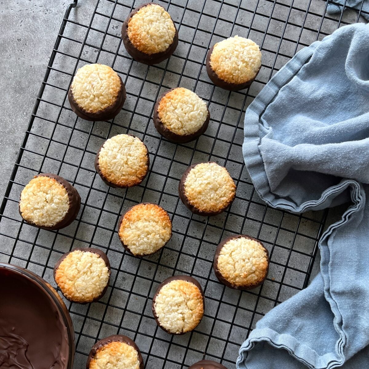 Coconut cookies dipped in chocolate, are arranged on a cooling rack next to a blue cloth and a bowl of melted chocolate.