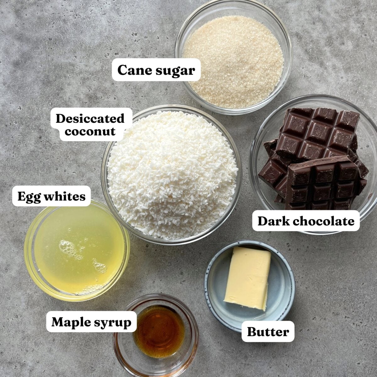 Six labeled bowls with ingredients for Coconut Cookies&mdash;cane sugar, desiccated coconut, dark chocolate, egg whites, maple syrup, and butter.