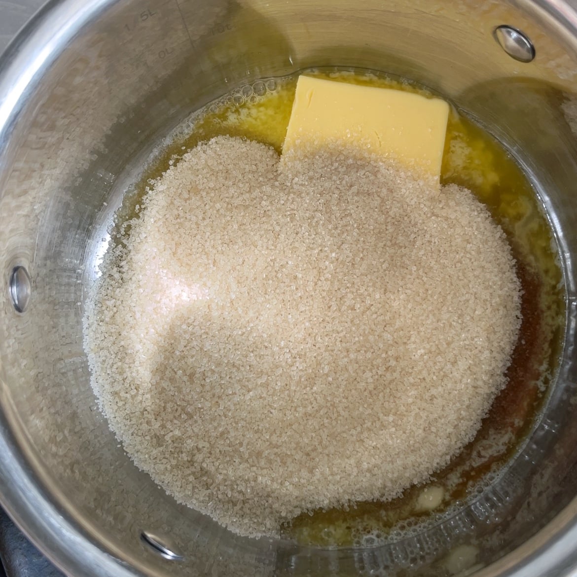 A metal pot containing butter, maple syrup, and a mound of granulated sugar.