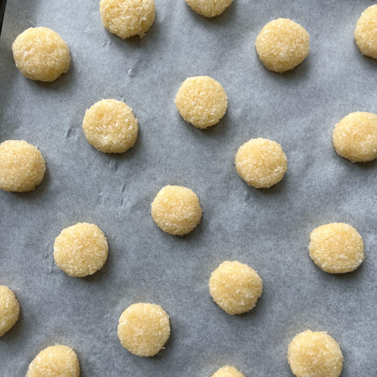 Balls of unbaked Coconut Cookies dough are arranged in rows on a parchment-lined baking sheet.