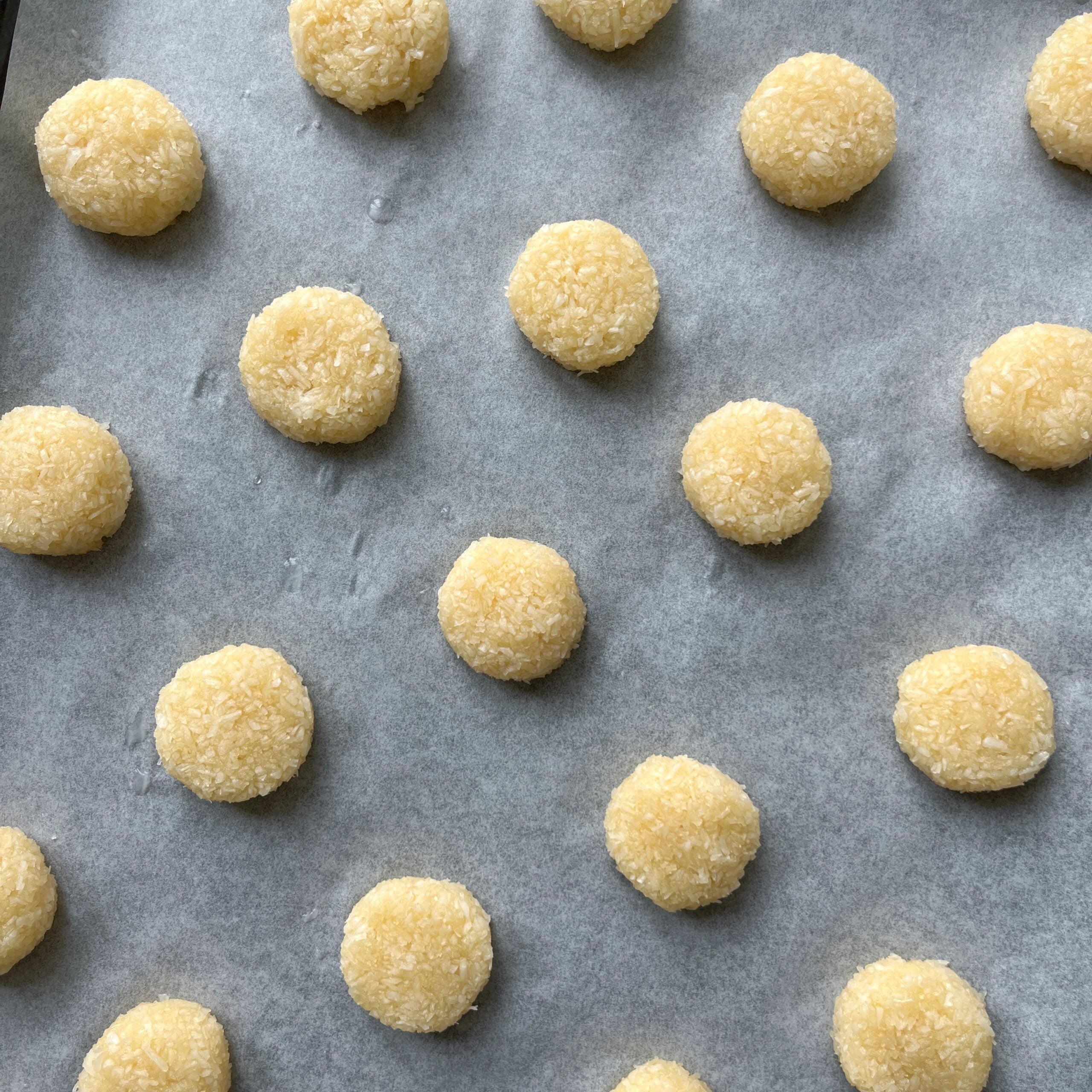 Balls of unbaked Coconut Cookies dough are arranged in rows on a parchment-lined baking sheet.
