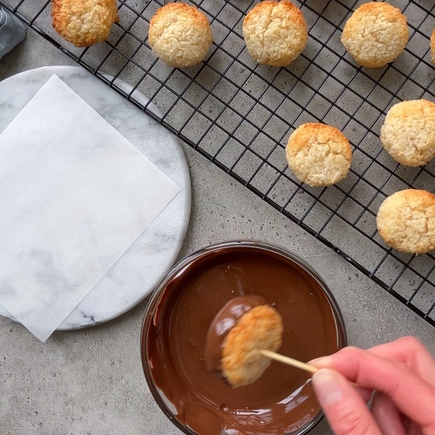 A hand dips a round baked coconut cookie into a bowl of melted chocolate, with more coconut cookies cooling on a wire rack nearby.