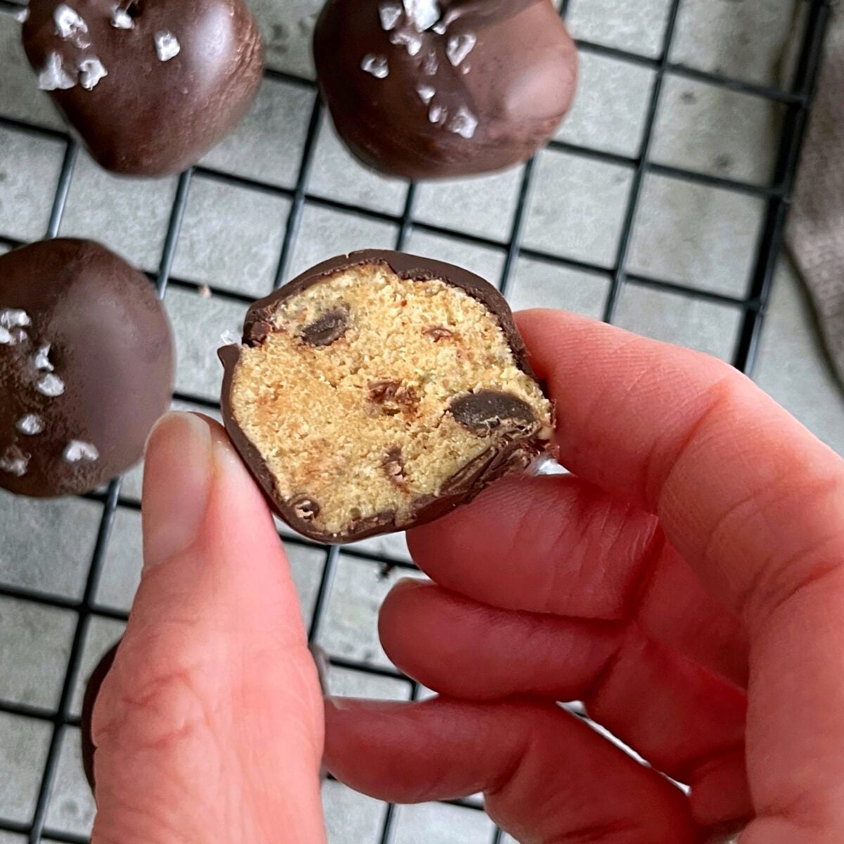 A hand holds a halved chocolate-coated Cookie Dough Bite with chocolate chips, above a cooling rack filled with more treats topped with sea salt.
