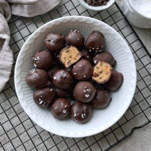 A white bowl filled with chocolate-covered cookie dough balls, some topped with flaky sea salt. One ball is cut open.