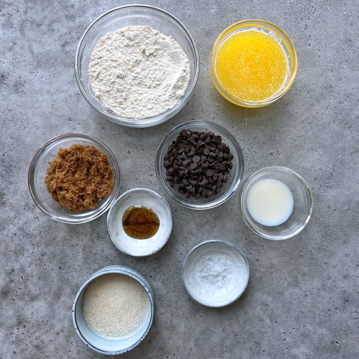 Eight bowls on a gray surface containing flour, melted butter, brown sugar, chocolate chips, milk, vanilla, sea salt, and cane sugar.