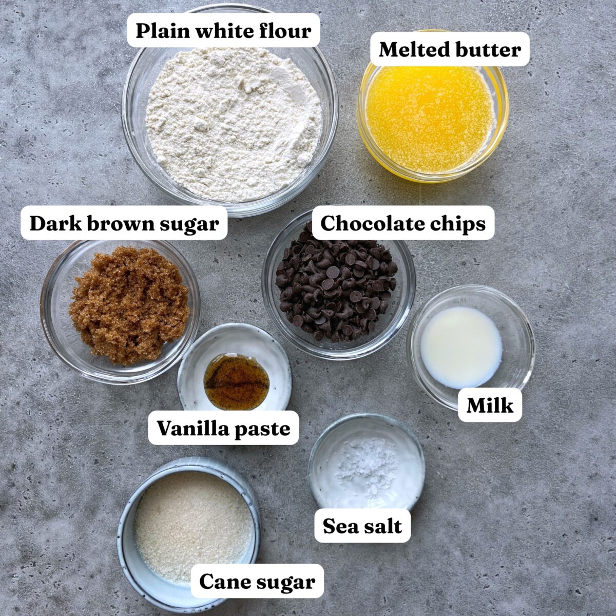 Eight labeled glass bowls containing plain white flour, melted butter, dark brown sugar, chocolate chips, vanilla paste, milk, sea salt, and cane sugar arranged on a gray surface.