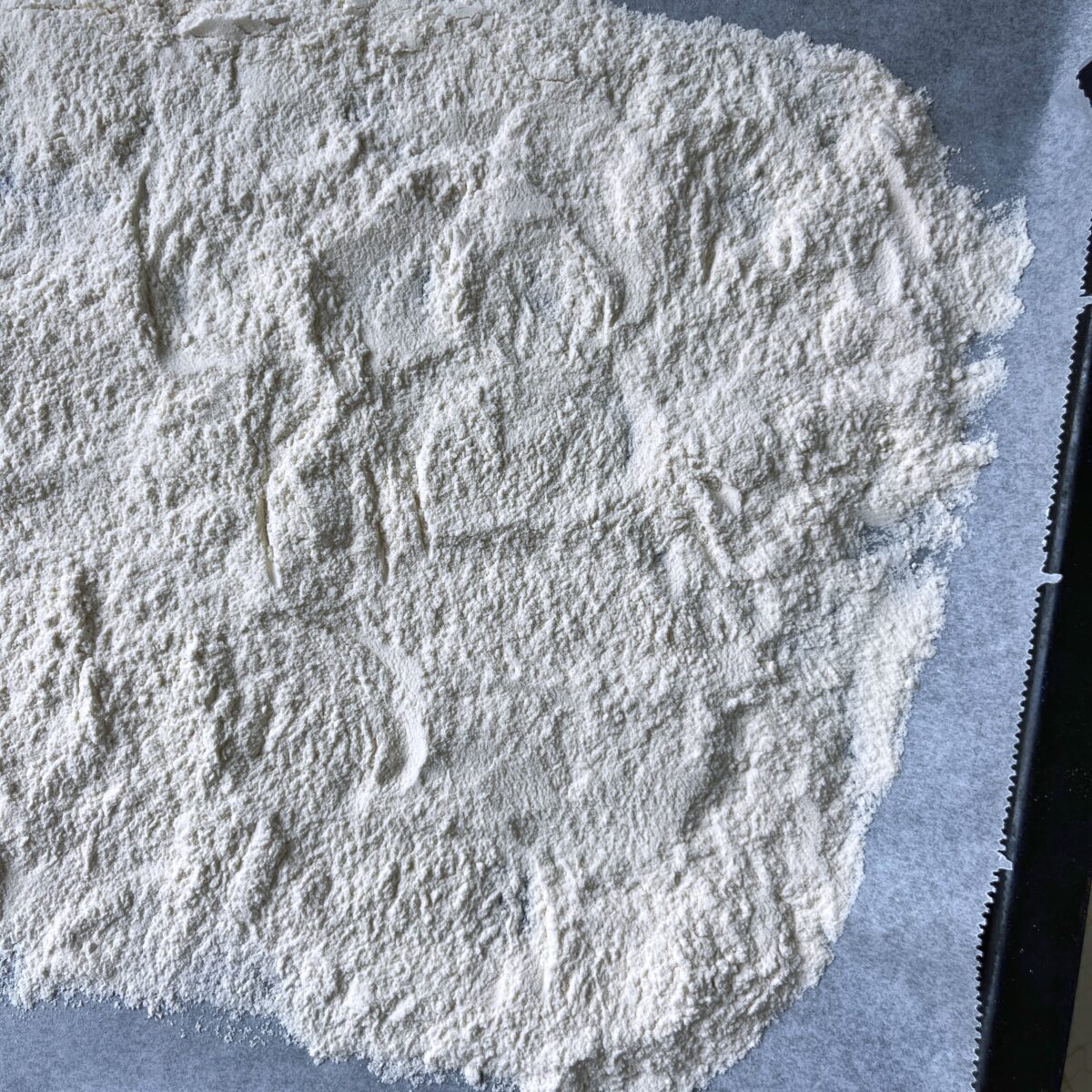 A layer of white flour spread evenly on a sheet of parchment paper, ready to heat-treat in the oven.