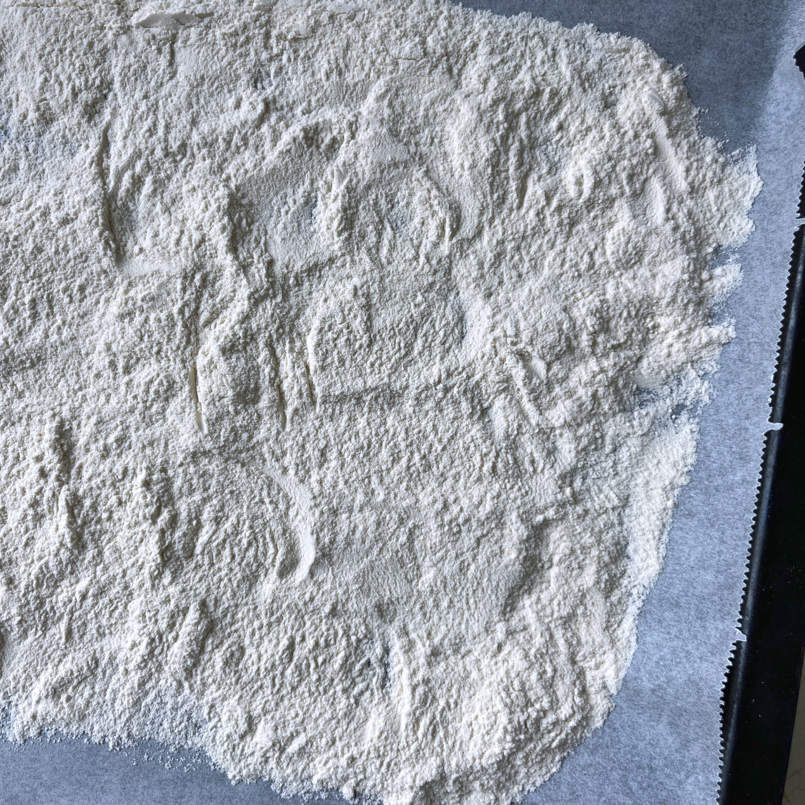 A layer of white flour spread evenly on a sheet of parchment paper, ready to heat-treat in the oven.