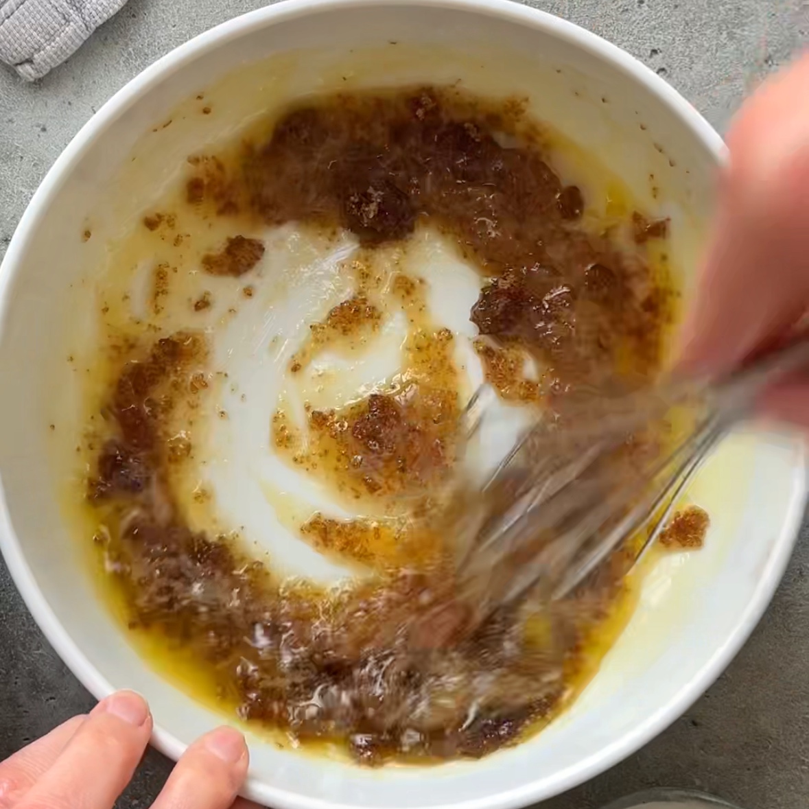 A person uses a whisk to mix brown sugar and cane sugar with melted butter in a white bowl on a gray countertop.