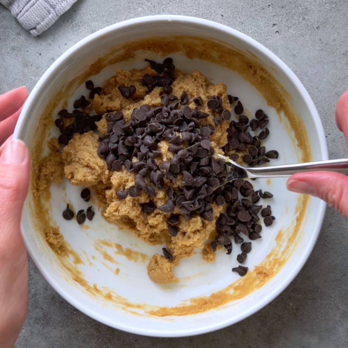 A person is mixing chocolate chips into cookie dough with a spoon in a white bowl on a gray surface.