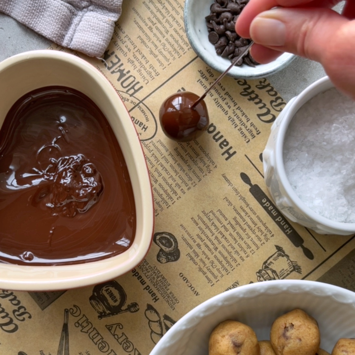A hand dips a Cookie Dough Bite into melted chocolate, with bowls of chocolate chips, salt, and cookie dough balls nearby on printed parchment paper.