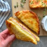A hand holds half of a toasted Croque Monsieur filled with ham and melted cheese, placed on a wooden board beside fresh herbs and a striped cloth napkin.