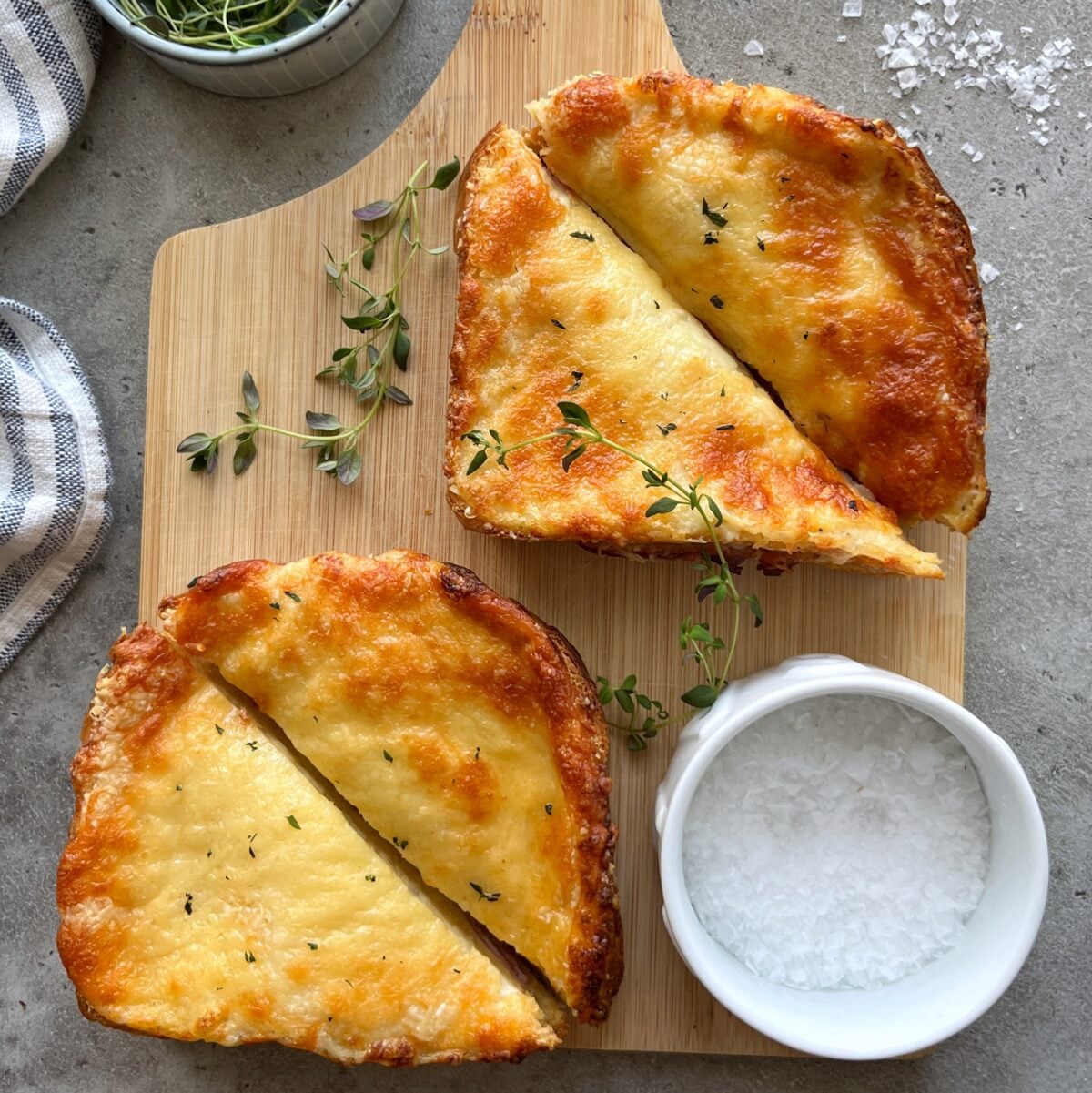 Two slices of cheesy Croque Monsieur toast cut in half, served on a wooden board with fresh herbs and a small bowl of coarse salt.