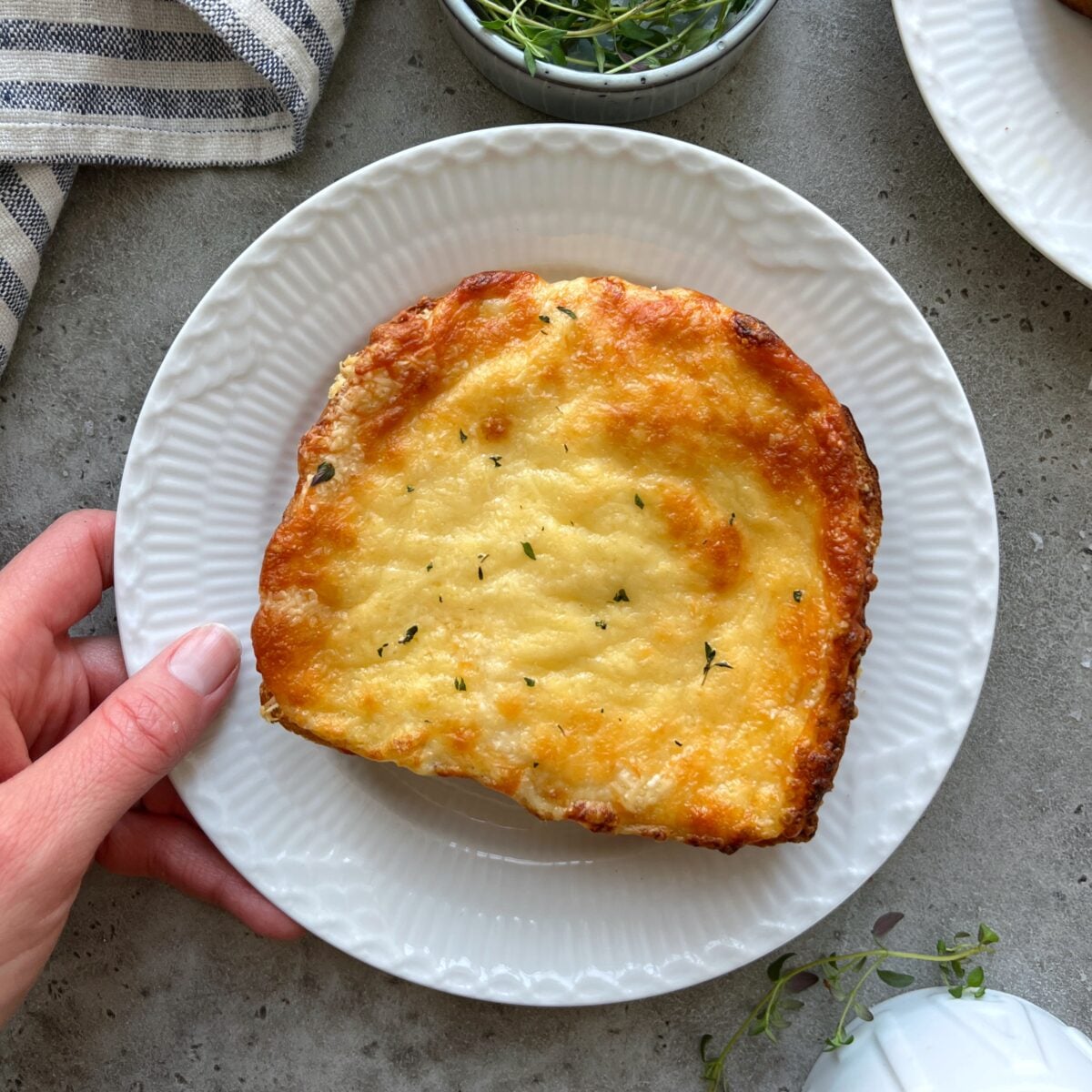 A hand holds a white plate with a classic Croque Monsieur.
