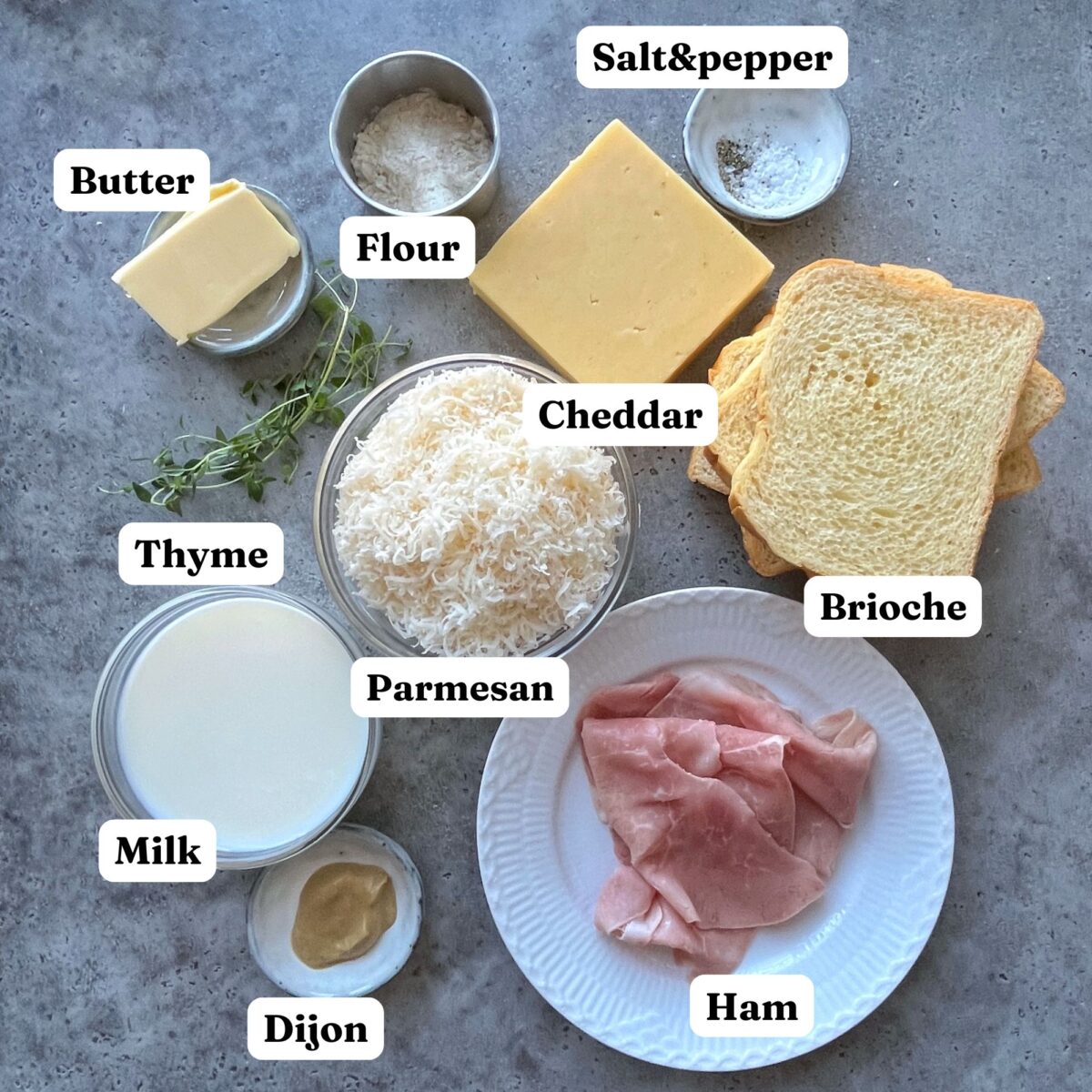 Ingredients for Croque Monsieur featuring sliced ham, brioche toast, cheddar, parmesan, butter, flour, milk, dijon mustard, thyme, salt, and pepper.