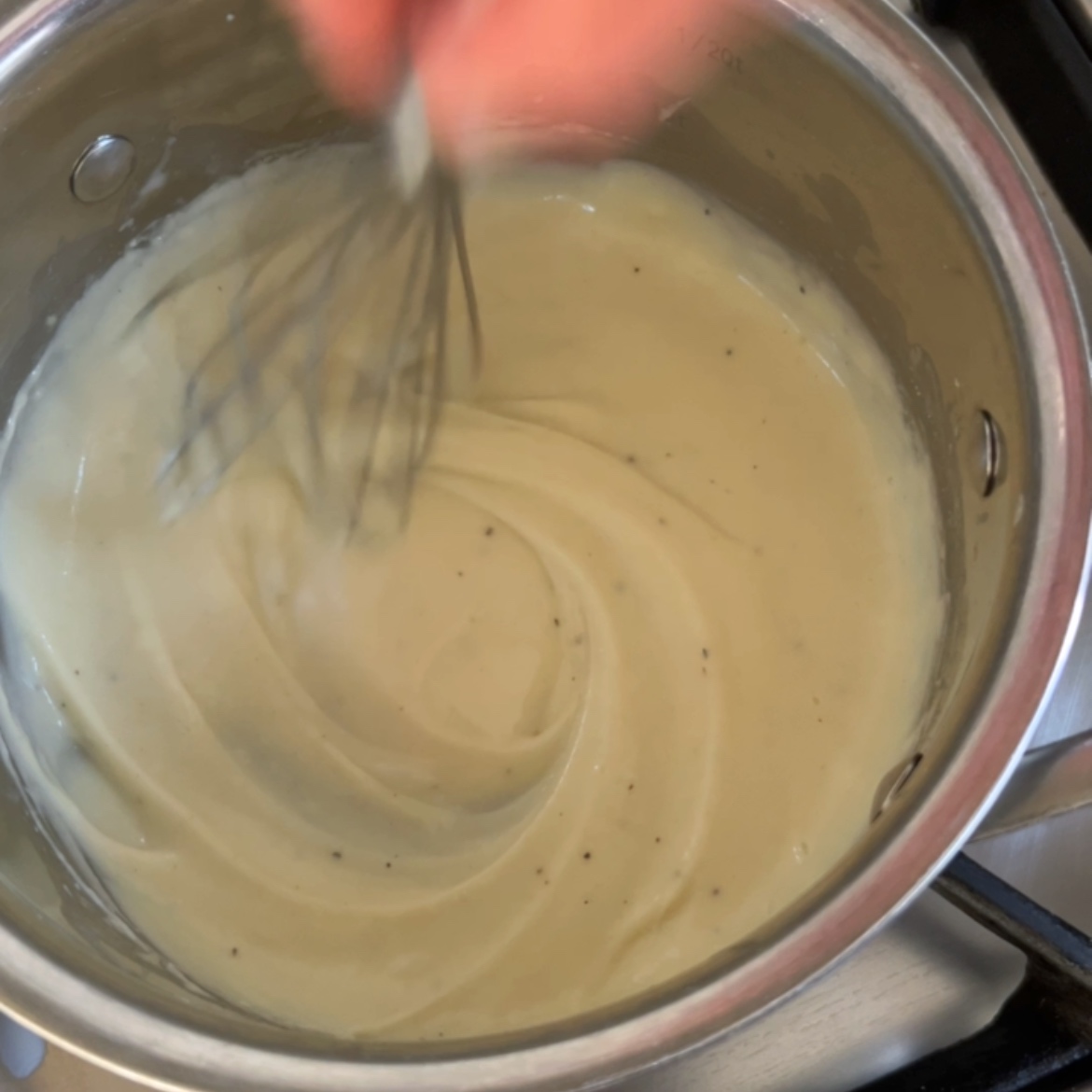 A person whisking a creamy b&eacute;chamel sauce in a stainless steel saucepan on a stove.