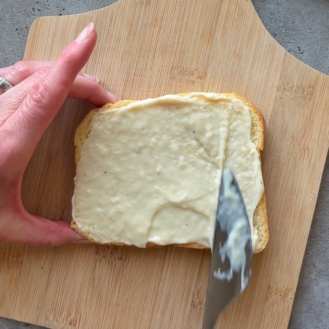 A hand spreads Bechamel onto a slice of brioche bread with a knife on a wooden cutting board.