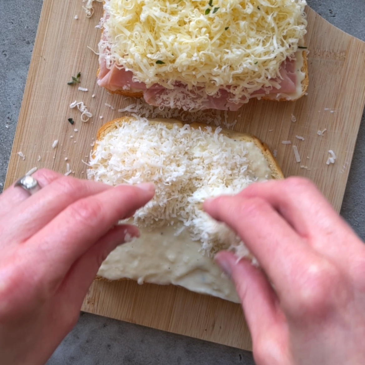 Two hands sprinkle shredded cheese onto a slice of bread with Bechamel, next to another slice topped with cheese and ham on a wooden cutting board.