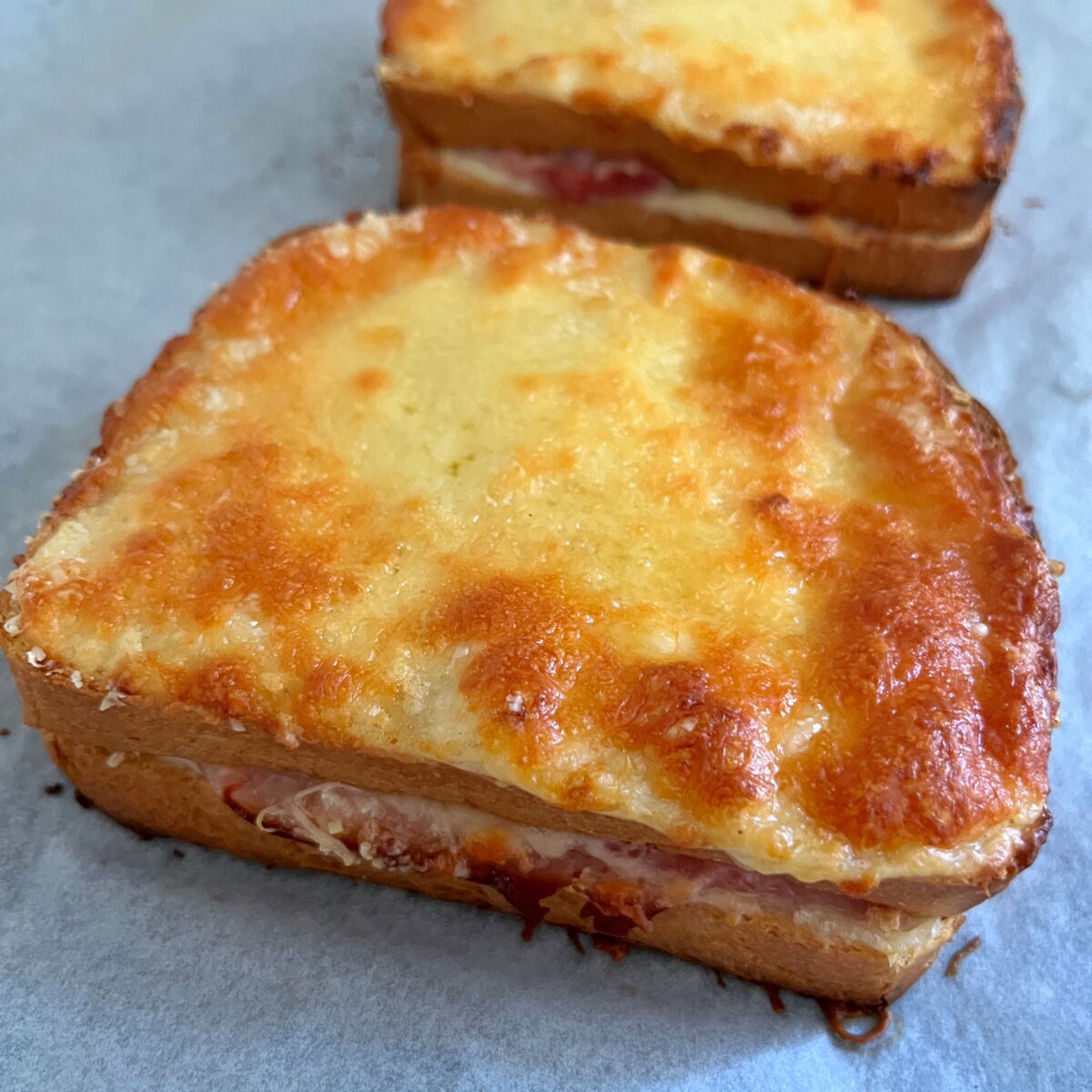 A toasted Croque Monsieur sandwich with melted cheese on top sits on parchment paper, with another similar sandwich in the background.