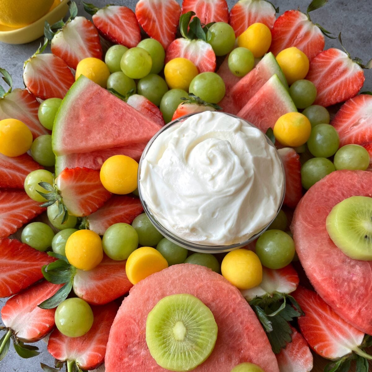 An Easter Fruit Tray featuring sliced strawberries, watermelon, green grapes, mango balls, and kiwi surrounds a bowl of lemon curd dip in the center.